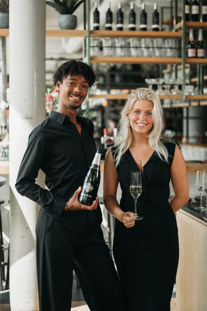 Twee representatieve hospitality professionals van Served With Passion B.V. verwelkomen gasten in een stijlvolle horecalocatie. De gastvrouw en gastheer dragen elegante zwarte kleding en stralen professionaliteit, gastvrijheid en klasse uit, perfect passend bij high-end events en luxe ontvangsten.