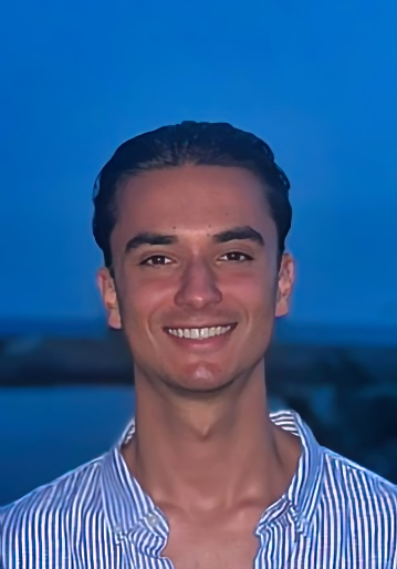 Portret van een jonge man in een gestreept overhemd met een brede glimlach, gefotografeerd bij zonsondergang aan het water. Deze foto hoort bij zijn referentie over zijn positieve ervaring bij Served With Passion, waarin hij het team beschrijft als liefdevol, energiek en gastgericht.