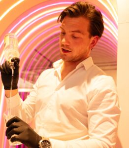 Man in een witte blouse en zwarte handschoenen schenkt een drankje in onder kleurrijke neonverlichting.