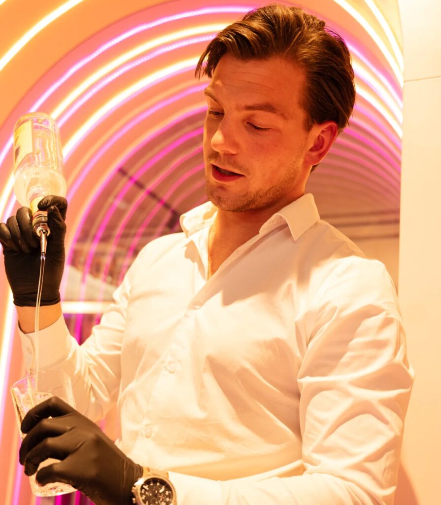 Man in een witte blouse en zwarte handschoenen schenkt een drankje in onder kleurrijke neonverlichting.