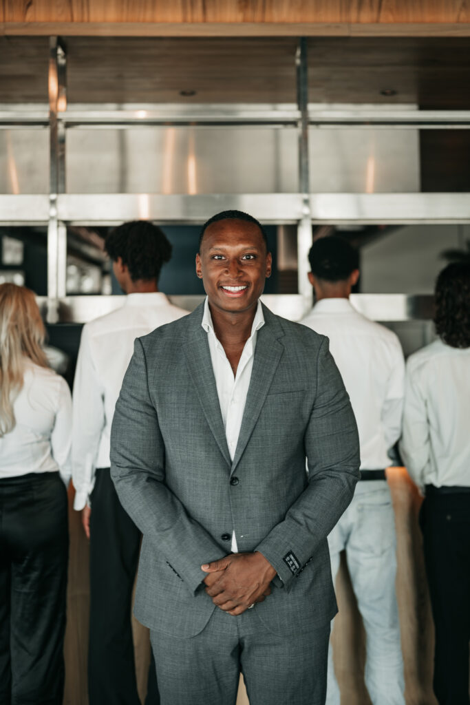 Supervisor van Served With Passion B.V. staat voor zijn team van hospitality professionals in een luxe horecalocatie. In een grijs pak en met een zelfverzekerde uitstraling vertegenwoordigt hij leiderschap, structuur en kwaliteit binnen de event- en horecabranche.