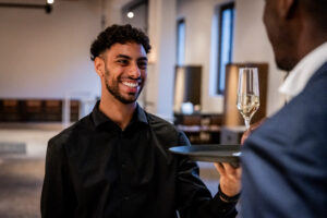Hospitalitymedewerker van Served With Passion glimlacht tijdens zijn werk terwijl hij een glas champagne serveert — symbool voor groei, zelfvertrouwen en doorgroeimogelijkheden binnen de horeca en evenementenbranche