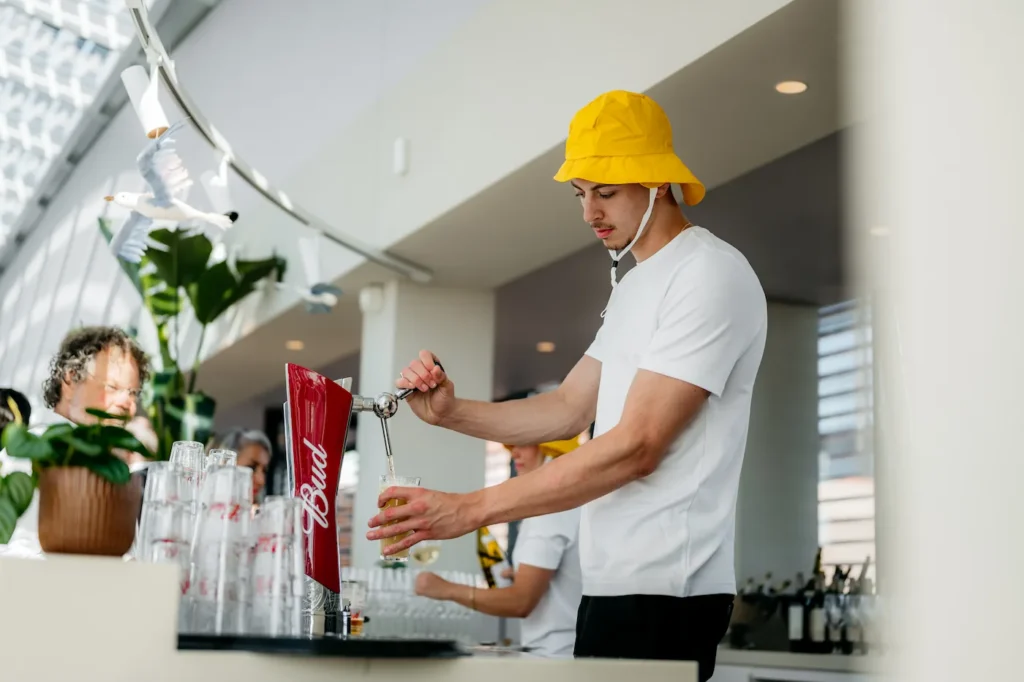 Man met gele hoed tapt een biertje uit een Bud-biertap in een moderne baromgeving.