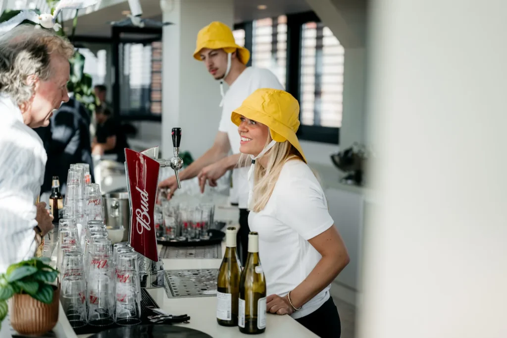 Twee medewerkers met gele vissershoeden tappen drankjes achter een bar tijdens een evenement.