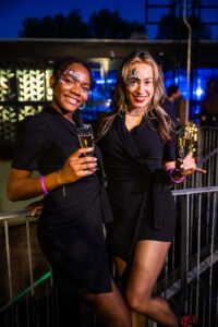 Twee vrouwen in zwarte outfits met gezichtsbeschildering poseren lachend met een glas champagne op een avondfeest.