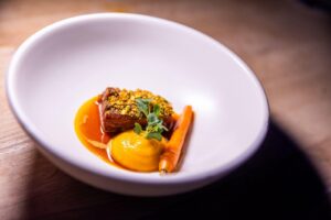 Fijn opgemaakt gerecht met een stuk vlees, wortelmousse, wortel en saus in een witte kom.