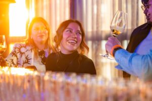 Drie mensen lachen en proosten met wijnglazen tijdens een sfeervol verlichte borrel.