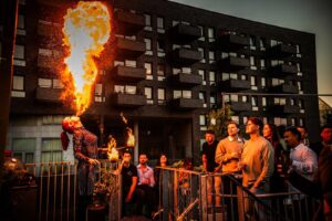 Vuurspuwer voert spectaculaire act uit voor een groep toeschouwers op een stadsterras bij zonsondergang.