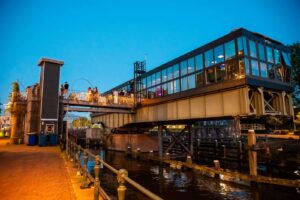 Avondfoto van restaurant WOLF, gevestigd in een oude spoorbrugconstructie boven het water met een verlicht terras vol mensen.
