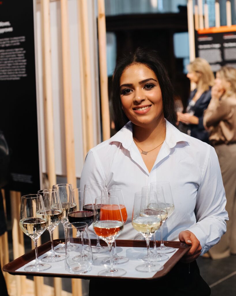 Portret van een lachende serveerster van Served With Passion tijdens een evenement. Ze draagt een witte blouse en houdt een dienblad met glazen wijn en frisdrank vast. De foto straalt gastvrijheid, professionaliteit en warmte uit — perfect passend bij de waarden van Served With Passion: enthousiasme, kwaliteit en passie voor hospitality