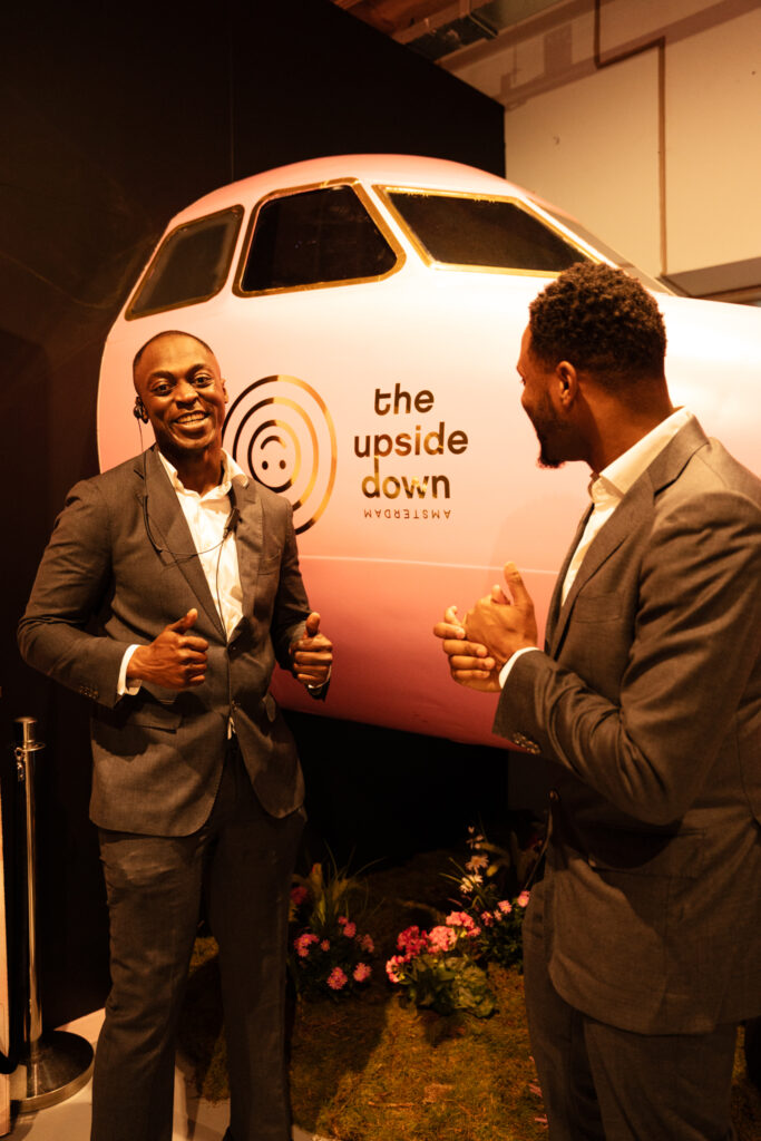 Twee vertegenwoordigers van Served With Passion B.V. tijdens het grootschalige buy-out event in The Upside Down Amsterdam. De heren, professioneel gekleed in grijze pakken, lachen terwijl ze voor de iconische roze vliegtuiginstallatie poseren — een moment dat de succesvolle samenwerking en energieke hospitality van Served With Passion weerspiegelt