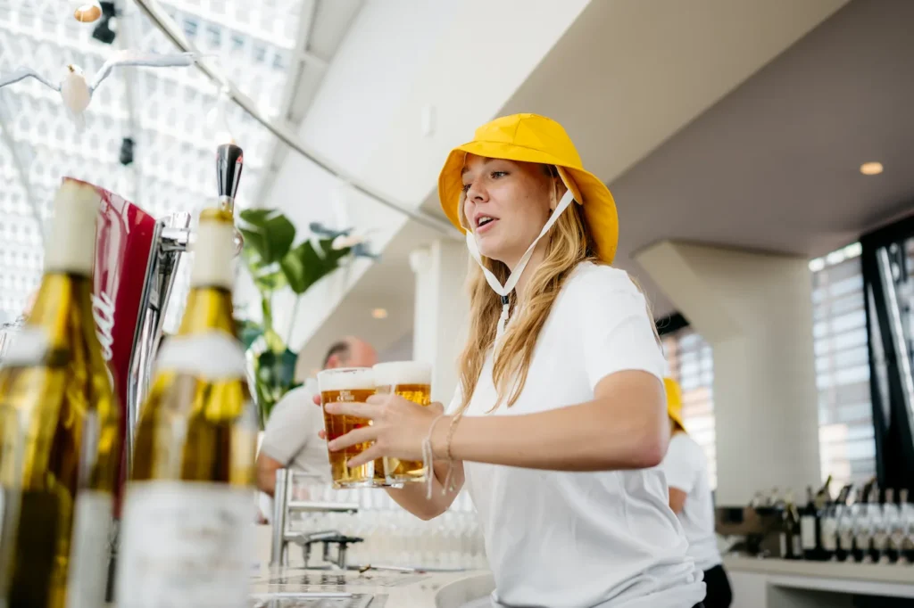 Vrouw met een gele hoed serveert bier aan de bar in een moderne, lichte ruimte.