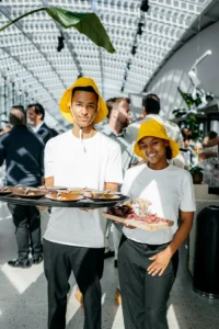 Twee jongvolwassenen in witte shirts en gele hoeden serveren hapjes tijdens een evenement in een moderne, lichte ruimte.