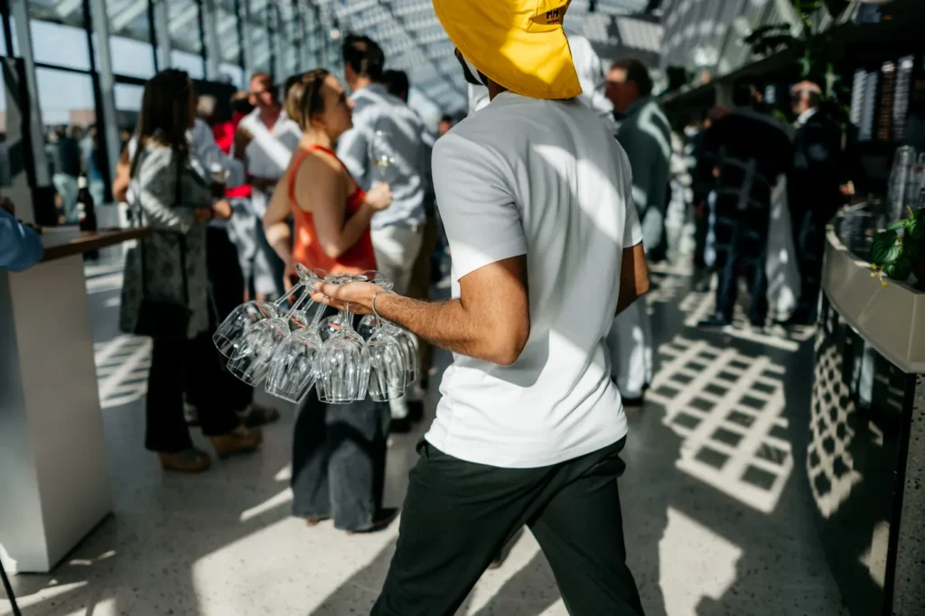 Een medewerker met een gele hoed draagt meerdere wijnglazen tijdens een drukbezocht evenement in een zonovergoten serre.