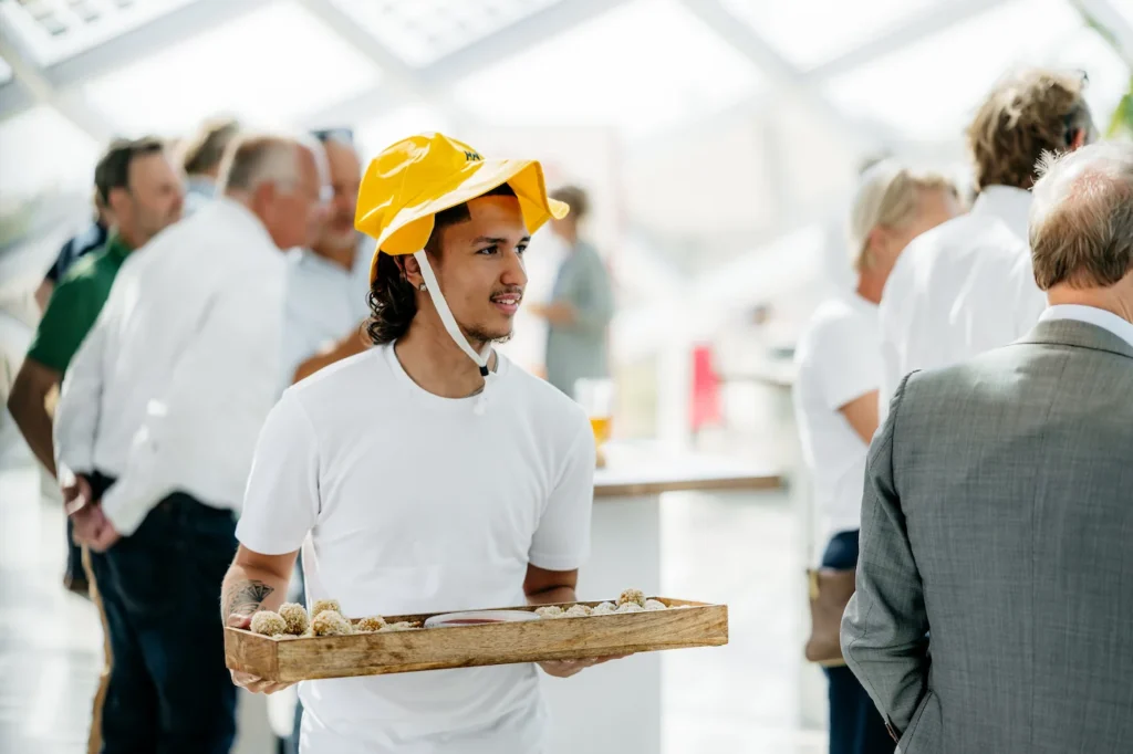 Man in wit shirt en gele hoed serveert hapjes tijdens een netwerkevenement.