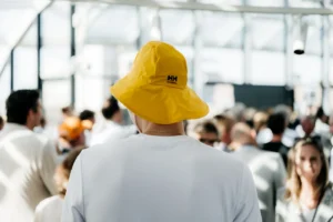 Man met gele HH-regenhoed kijkt uit over een drukke menigte in een lichte, overdekte ruimte.
