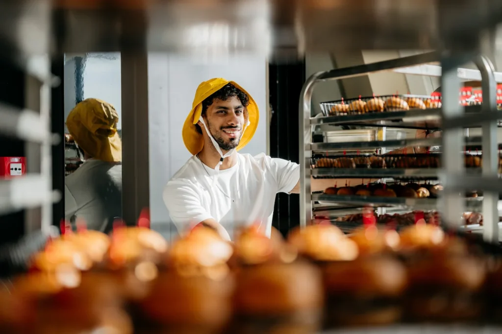 Man met een gele pet plaatst een rek vol versgebakken broodjes in een bakkerij.