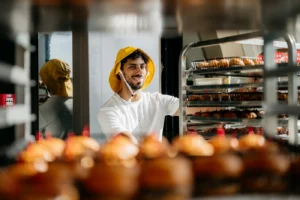 Man met een gele pet plaatst een rek vol versgebakken broodjes in een bakkerij.