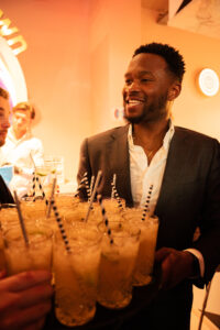 Een man in pak ontvangt lachend een dienblad met cocktails tijdens een evenement.