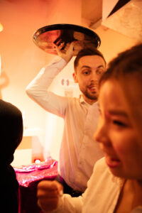 Man met serveerschaal boven zijn hoofd tijdens een levendig feestmoment met anderen op de voorgrond.