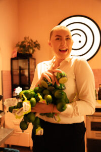 Vrolijke vrouw in witte blouse houdt meerdere netzakken limoenen vast in een warme, sfeervolle keuken.