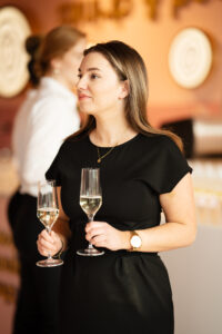Vrouw in een zwarte jurk met twee glazen champagne tijdens een evenement.