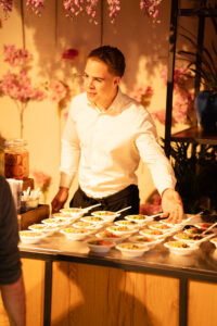 Een gastheer in wit overhemd serveert kleine kommetjes eten vanaf een sfeervol versierde foodstand met roze bloemen.