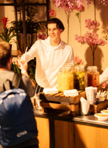 Een vriendelijke medewerker in wit overhemd overhandigt een drankje aan een bezoeker bij een kleurrijk gedecoreerde drankentafel.