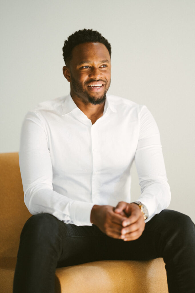Denzell Denneboom, oprichter en directeur van Served With Passion B.V., zittend op een beige stoel in een lichte ruimte. Zijn glimlach en uitstraling weerspiegelen de kernwaarden van Served With Passion: professionaliteit, warmte en toewijding aan hoogwaardige hospitality.