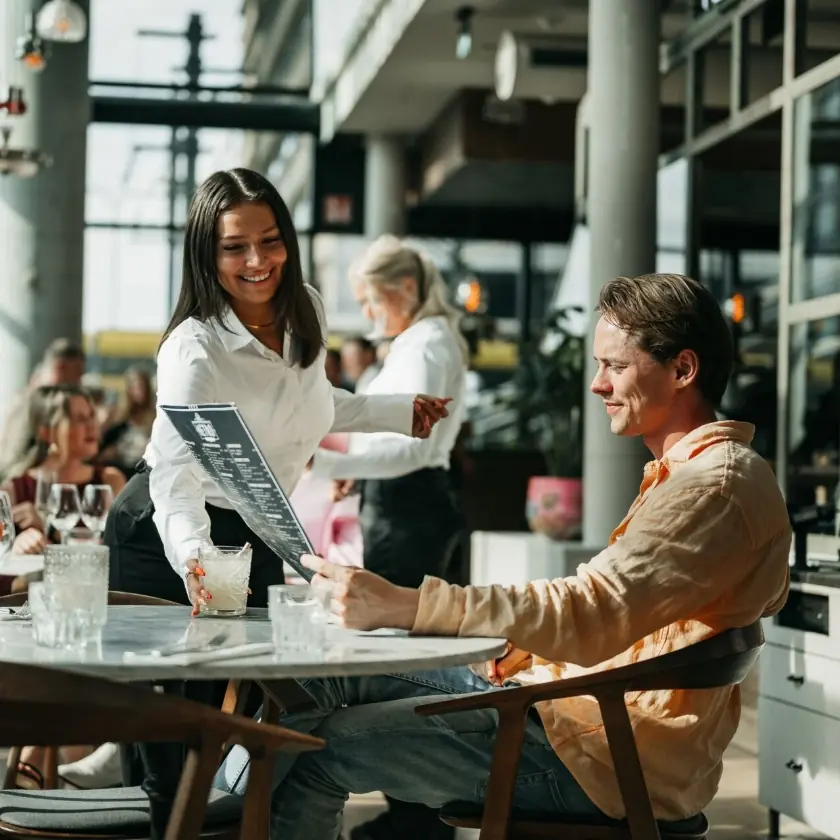 Serveerster overhandigt een drankje en menukaart aan een mannelijke gast in een modern restaurant.