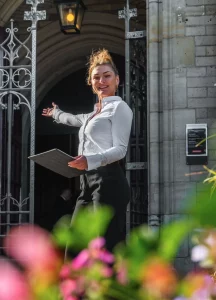 Gastvrouw verwelkomt bezoekers bij een entree met sierlijk hekwerk en historische architectuur.