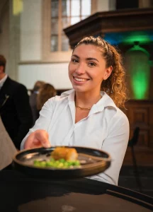 Vriendelijke serveerster in witte blouse presenteert een gerecht op een zwart bord tijdens een hospitality-evenement.