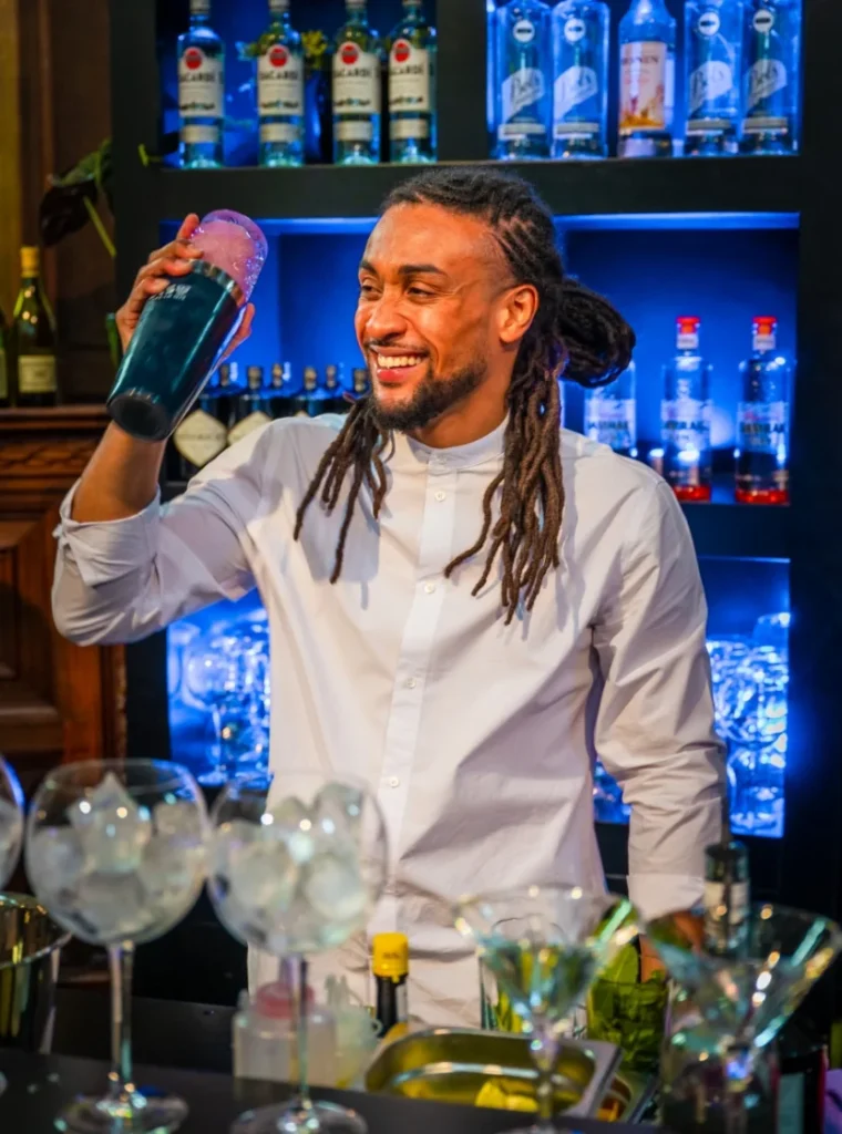 Barman in wit overhemd maakt een cocktail met een shaker achter een bar vol drankflessen en glazen.