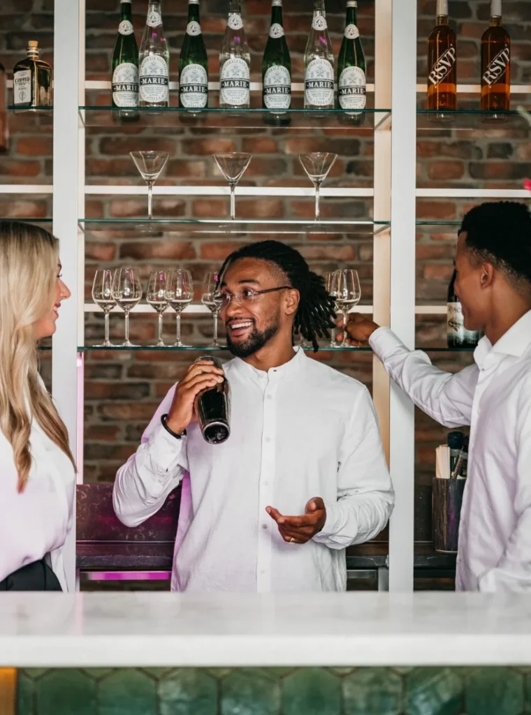 Drie bartenders in witte overhemden praten en lachen achter een stijlvolle bar met flessen en glazen op de achtergrond.