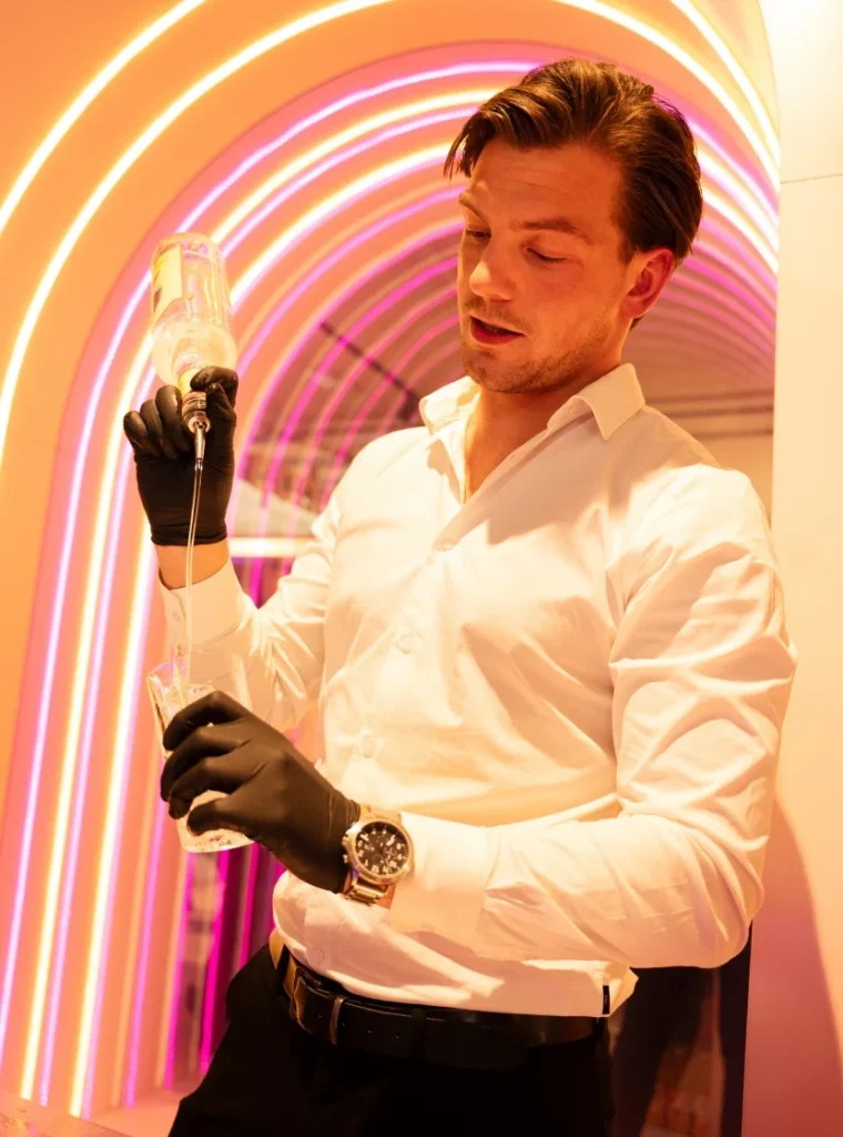 Barman in witte blouse en zwarte handschoenen schenkt drank in een glas onder kleurrijke neonverlichting.
