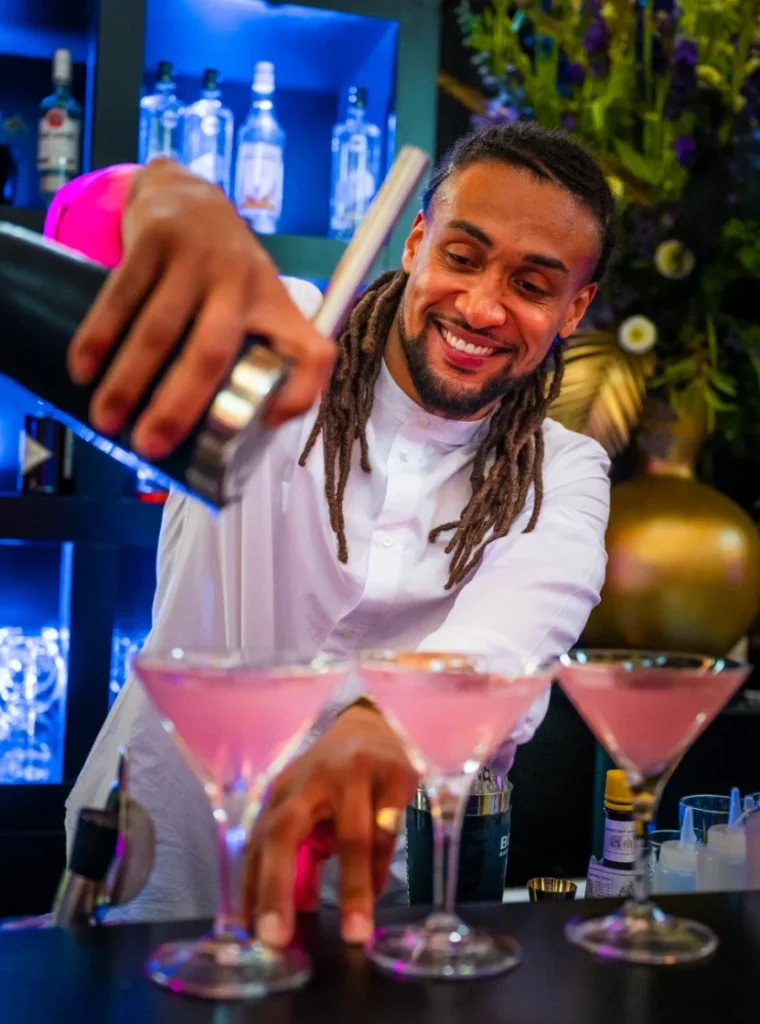 Een glimlachende bartender schenkt roze cocktails in martiniglazen achter een stijlvolle bar.