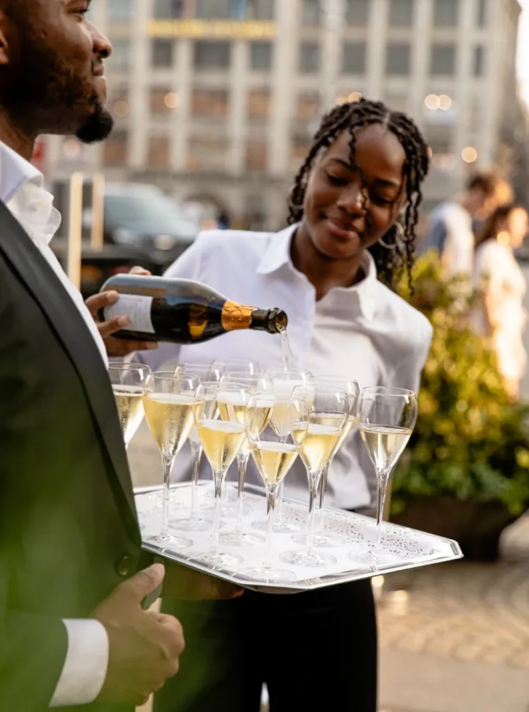 Twee obers in witte overhemden schenken champagneglazen vol tijdens een buitenevenement.