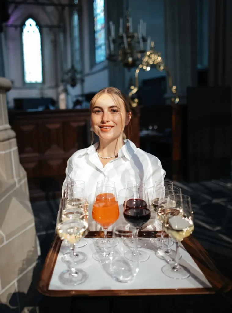 Vriendelijke serveerster met een dienblad vol diverse drankjes in een sfeervolle kerkelijke omgeving.