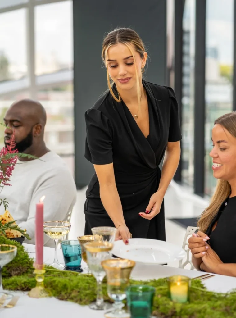 Een vrouwelijke hostess in zwart jurkje serveert borden aan een feestelijk gedekte tafel tijdens een evenement.
