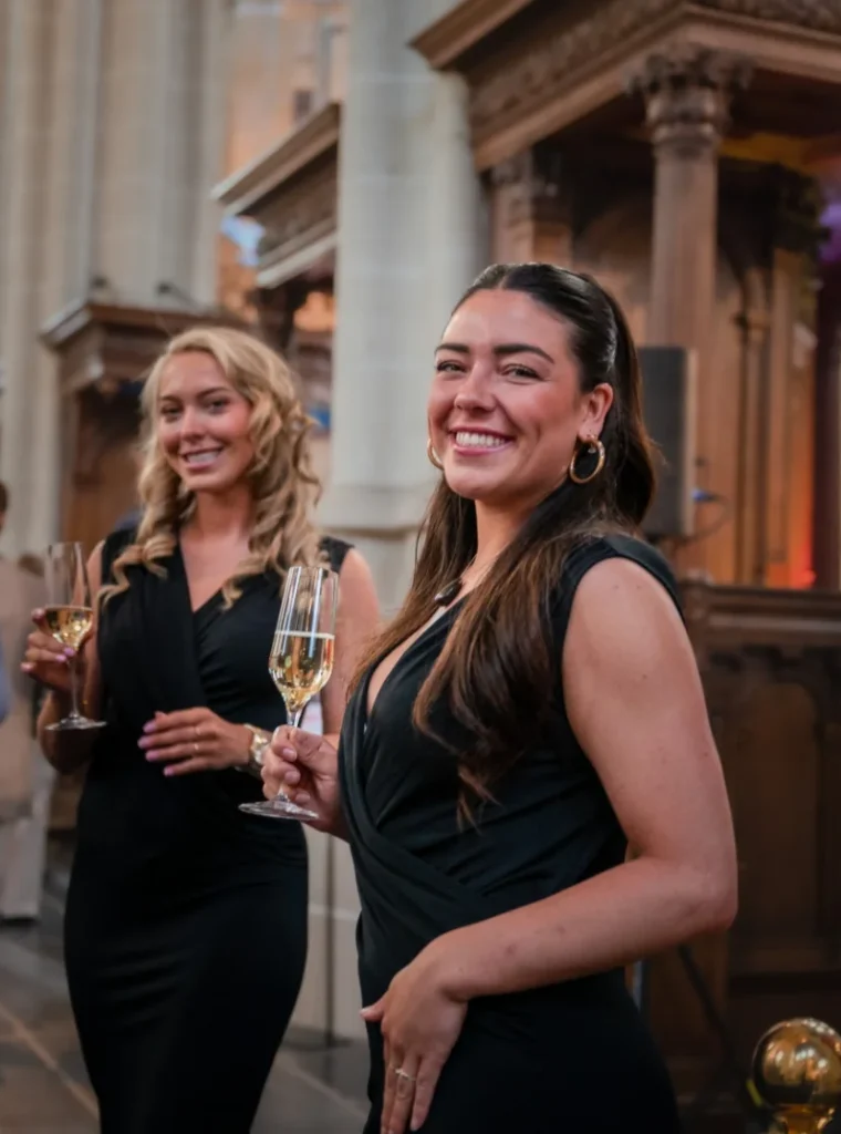 Twee hostessen in elegante zwarte jurken poseren met een glas champagne in een sfeervolle locatie.