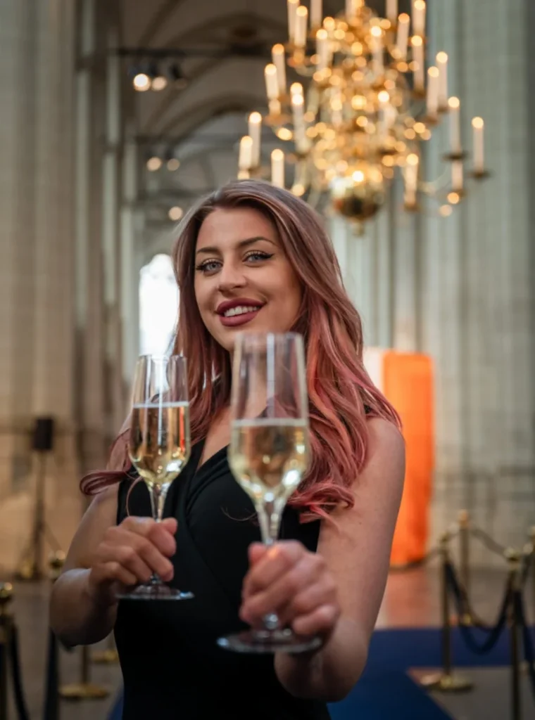 Hostess met twee glazen champagne in een feestelijke, chique setting met kroonluchter op de achtergrond.