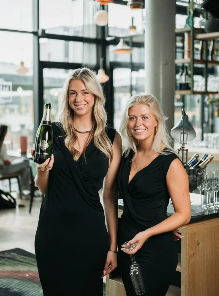 Twee hostessen in zwarte jurken poseren met een fles champagne en een glazen karaf in een moderne bar.