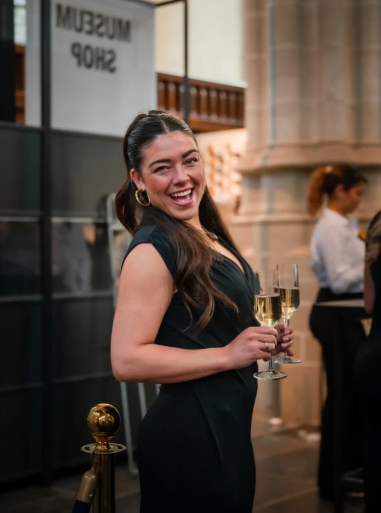 Vrouwelijke host in zwarte jurk lacht terwijl ze twee glazen champagne vasthoudt tijdens een evenement in een museum.