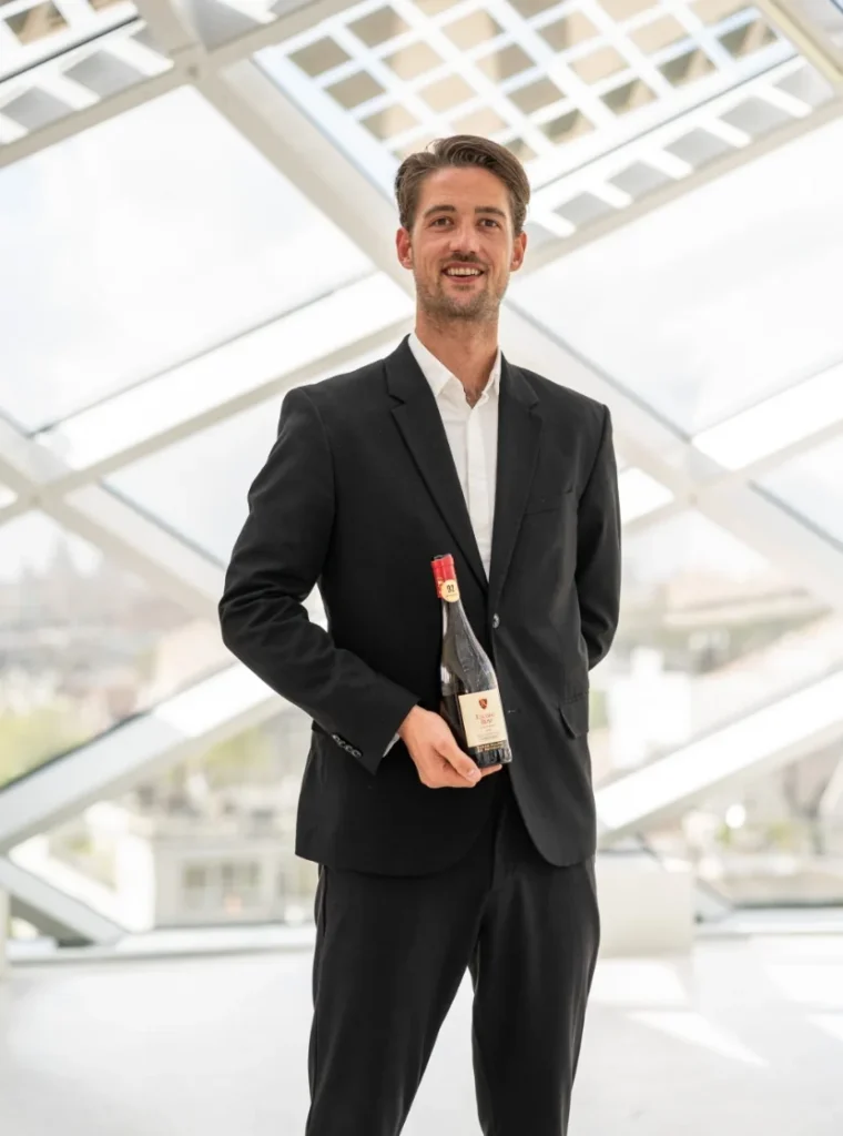 Man in pak poseert met fles wijn in moderne, lichte ruimte.