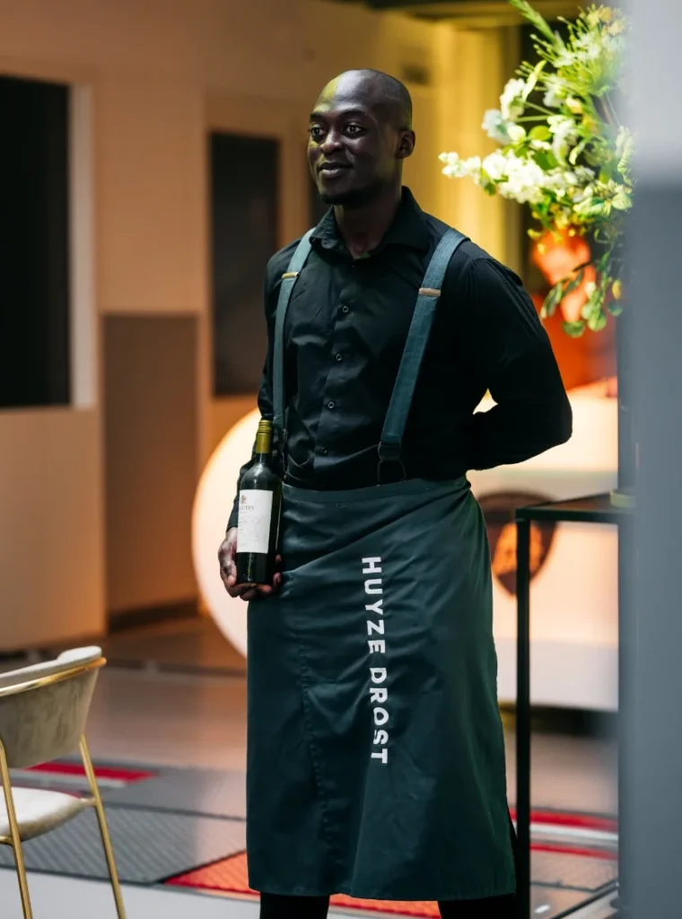 Ober van Huize Drost in zwart uniform met wijnfles in de hand tijdens een evenement.