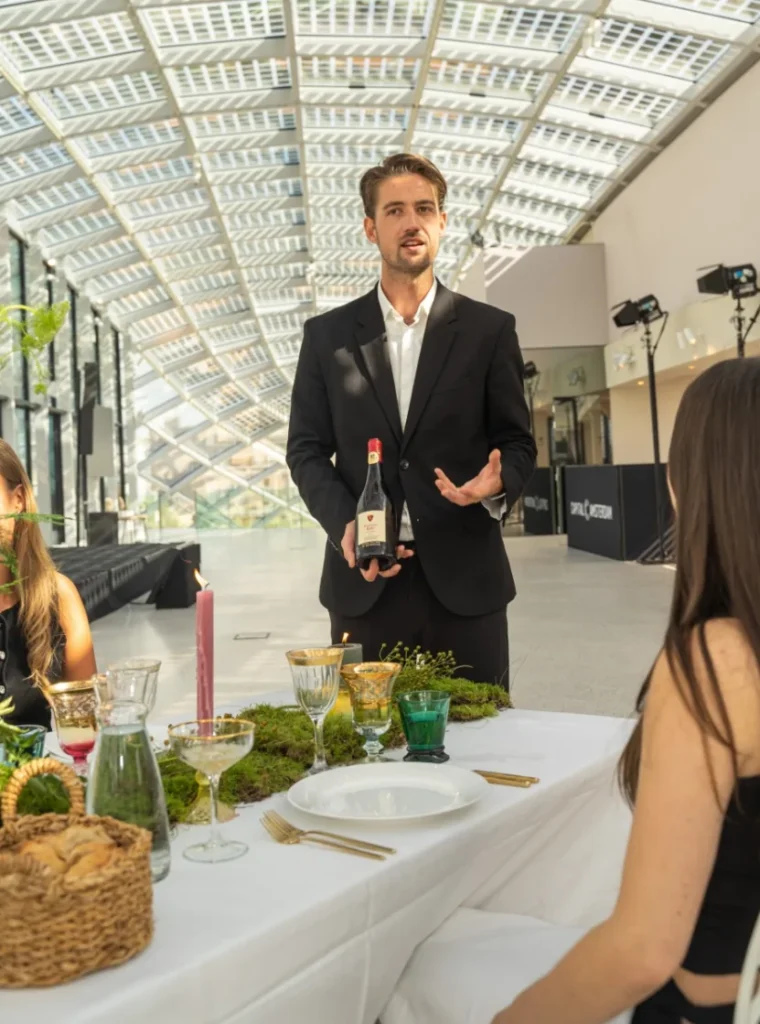 Man in pak presenteert een fles wijn aan een gedekte tafel met gasten in een moderne, glazen hal.