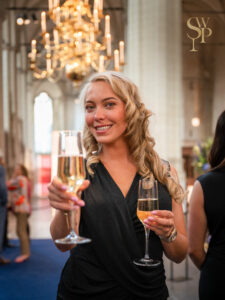 Vrouw in een elegante zwarte jurk houdt twee glazen champagne vast tijdens een chique evenement in een sfeervol verlichte zaal.