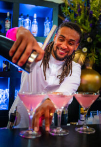 Barman schenkt roze cocktails in martini glazen achter een verlichte bar.