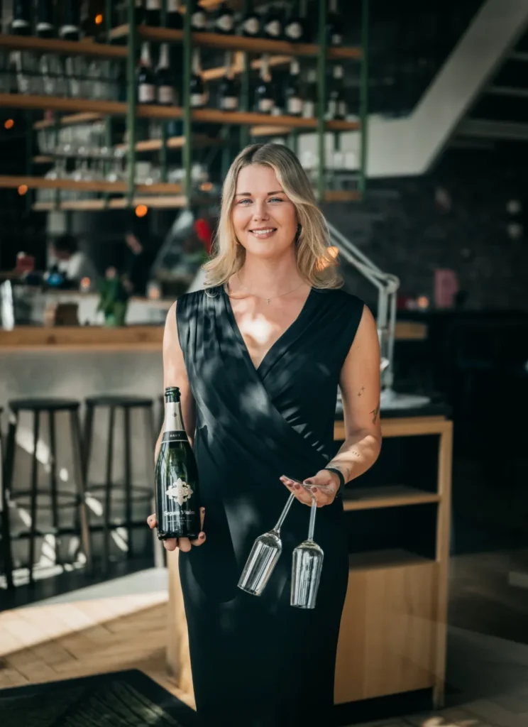 Naomi van Nieuwland in een zwarte jurk met een fles mousserende wijn en twee glazen in een moderne bar.