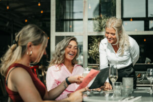 Served With Passion-medewerkster in gesprek met gasten op een zonnig terras, terwijl ze met een glimlach helpt bij het kiezen van een drankje. Deze foto symboliseert de persoonlijke benadering van Served With Passion: niet uitzenden, maar écht matchen tussen mens en merk.